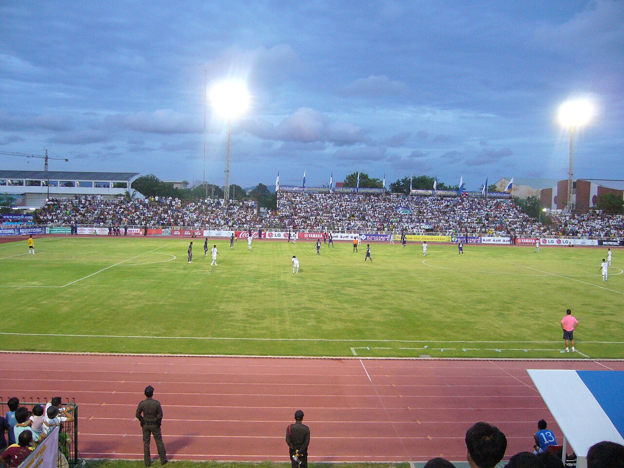 A packed crowd watches a match at Rayong Province Stadium under floodlights, with a running track in the foreground