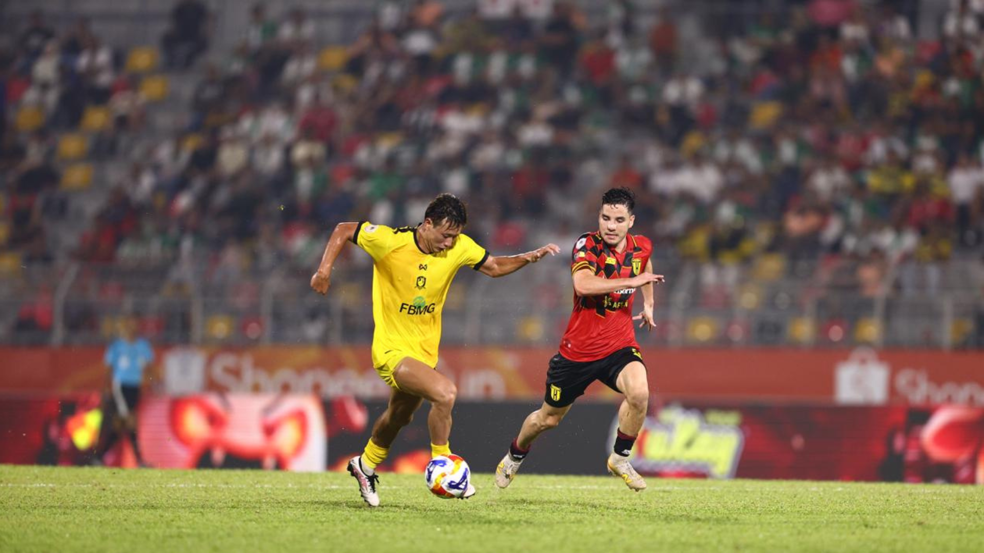 A Kuching City player in yellow runs with the ball, chased by a Tampines Rovers player in red and black